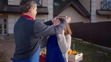 Loving couple putting on aprons standing on front yard selling organic products. Positive confident Caucasian husband and wife with healthful fruits and vegetables outdoors. Farming and lifestyle
