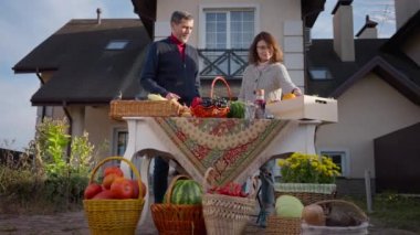 Wide shot happy farmers talking smiling standing at table with organic fruits and vegetables on front yard. Positive loving adult Caucasian couple of gardeners selling local products. Slow motion