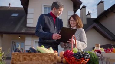 Medium shot portrait of laughing Caucasian woman talking with positive adult man standing at table with organic local products. Happy confident couple of farmers selling fruits and vegetables outdoors