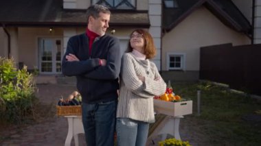 Confident positive husband and wife posing back to back on front yard selling local organic fruits and vegetables. Portrait of smiling Caucasian adult couple looking at camera with hands crossed
