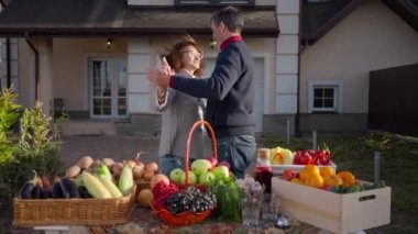 Loving couple of gardeners dancing at table with organic local products. Live camera zoom in and zoom out as happy Caucasian husband and wife enjoying sunny autumn day selling fruits and vegetables