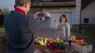 Back view man taking photo of woman posing with organic local products on farm market. Shooting over shoulder of loving husband supporting wife selling healthful fruits and vegetables