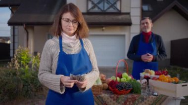 Portrait of Caucasian adult woman counting cash income posing standing on the left with blurred man selling local fruits and vegetables at background. Couple of farmers outdoors on autumn day