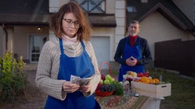Concentrated Caucasian woman counting cash standing on farm market with blurred man in apron at background. Happy satisfied wife and husband selling local healthful products outdoors. Slow motion