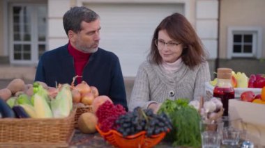Front view of thoughtful Caucasian adult couple talking sitting at table with local organic products. Male and female farmers selling fruits and vegetables chatting on farm market