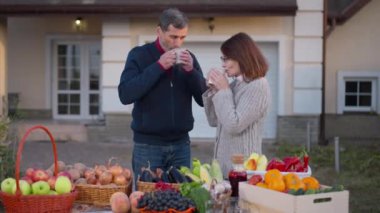 Caucasian husband and wife drinking morning coffee standing on farm market with organic products on table. Happy confident couple of farmers talking enjoying morning outdoors on autumn day