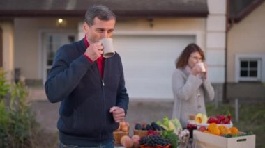 Male farmer drinking morning coffee looking back at blurred woman enjoying hot drink at background. Portrait of positive Caucasian husband and wife on farm market outdoors