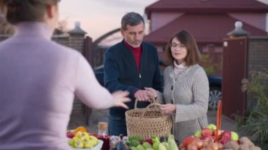 Adult couple choosing vegetables on farm market with seller talking advertising organic products. Positive confident Caucasian husband and wife buying veggies outdoors in the evening