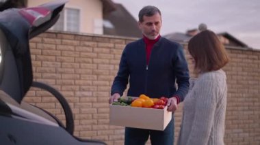 Satisfied couple talking putting box of organic healthful vegetables in car trunk. Happy smiling Caucasian man and woman buying local products on farm market in the evening. Lifestyle concept