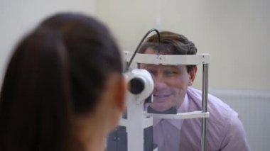 Zoom in to smiling Caucasian man sitting at slit lamp in hospital as doctor examining eye. Positive confident patient on appointment with professional ophthalmologist indoors
