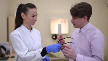 Thankful Caucasian man receiving eyeglasses from doctor looking at camera smiling. Portrait of grateful patient and intelligent Caucasian doctor standing in ophthalmological clinic