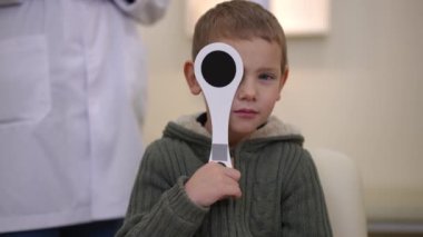Portrait of cute charming Caucasian boy closing one eye with eye occluder reading talking. Front view of child undergoing medical examination passing eye test in hospital indoors