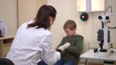 Back view woman examining painful eye of blurred little patient in ophthalmological clinic. Shooting over shoulder of professional Caucasian woman calming down child in hospital