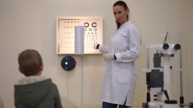 Beautiful positive doctor standing at eye sight test chart pointing listening to boy talking. Portrait of expert Caucasian woman doing medical checkup for little patient in ophthalmological hospital