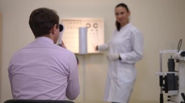 Back view unrecognizable man undergoing eyesight test gesturing sitting on the left with blurred expert doctor at background. Caucasian patient and professional woman in ophthalmological hospital