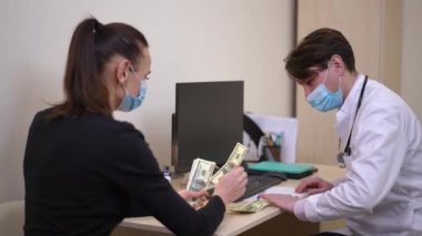 Portrait of unsure doctor in coronavirus face mask trying refuse bribe as woman putting money on table. Caucasian man receiving cash from patient in hospital. Bribery and medicine concept