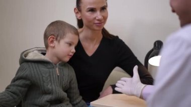 Smiling mother talking with doctor as man gesturing high-five with little boy. Positive satisfied Caucasian woman with child on appointment in hospital indoors