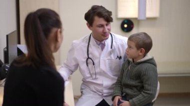 Charming boy sitting on lap of smiling doctor answering questions with blurred woman at front. Positive Caucasian man asking little patient filling in medical history smiling. Pediatrics concept