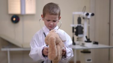 Front view portrait of cute Caucasian little boy in white medical gown with stethoscope playing with toy. Medium shot of curios child in hospital indoors. Medicine and future occupation concept