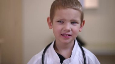 Live camera zoom out from thoughtful Caucasian little boy posing in hospital wearing white gown. Unsure hesitating child choosing dream job profession thinking looking away