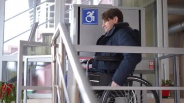 Side view man rolling wheelchair from lift platform leaving building. Confident Caucasian ill guy riding mobility aid device with hands. Accessibility concept