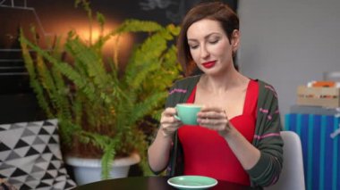 Medium shot portrait of gorgeous confident woman tasting delicious coffee looking away smiling. Beautiful Caucasian female client enjoying freshly brewed hot drink in cafe indoors