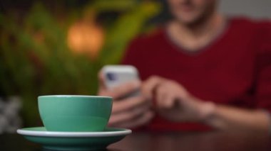 Green coffee cup on table in cafe with blurred Caucasian man scrolling social media on smartphone at background. Unrecognizable male client surfing Internet messaging online in coffee shop indoors