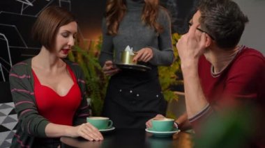 Adult couple talking at table in cafe as unrecognizable waitress bringing bill taking away coffee cups. Confident Caucasian man and woman dining having lunch in coffee shop indoors