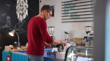 Caucasian man taking straw and sugar on cafe counter leaving with takeaway cup. Confident male client in coffee house indoors walking away with purchase. Self service. Coffee names in Russian on wall