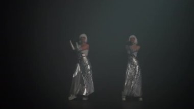 Wide shot two professional dancers in stage costumes dancing in foggy darkness. Portrait of slim Caucasian beautiful women performing at black background in mist. Art and performance concept