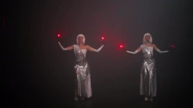 Two women in cyber outfit dancing spinning turning on red neon light on gloves performing in fog on stage. Wide shot portrait of talented performers in futuristic dresses dancing with laser beam