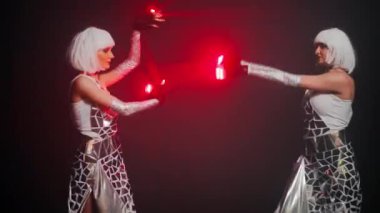 Medium shot modern dance of two confident talented Caucasian women in futuristic dresses with red laser light. Portrait of professional dancers looking at camera performing on stage posing