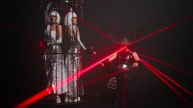 Wide shot women in stage dresses dancing in cage and man in costume with red laser light sitting on metal chair. Caucasian dancers performing contemporary dance on stage in darkness. Slow motion
