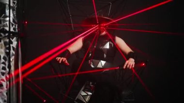 Man in futuristic samurai costume sitting on metal chair looking around as women dancing in cage on stage. Zoom in to Caucasian performer at black background. Slow motion