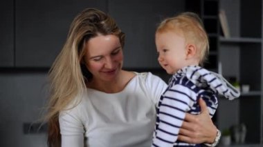 Smiling happy Caucasian mother holding baby daughter in hands talking kissing toddler. Portrait of beautiful woman enjoying leisure with infant standing in kitchen at home indoors
