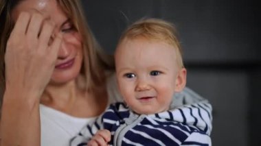 Close-up cute blond baby girl with grey eyes looking at camera smiling as woman kissing child on cheek. Portrait of curios positive happy daughter and loving mother indoors at home