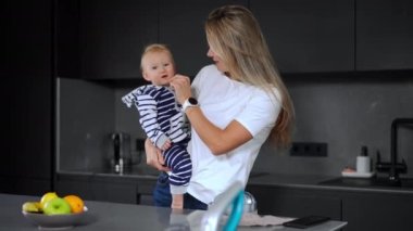 Beautiful woman caressing infant face with feather looking at camera smiling. Portrait of happy loving Caucasian mother playing with baby daughter standing in kitchen at home indoors posing