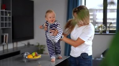 Joyful charming little girl doing first steps with happy mother holding baby. Smiling curios Caucasian infant daughter enjoying learning walking with parent at home indoors