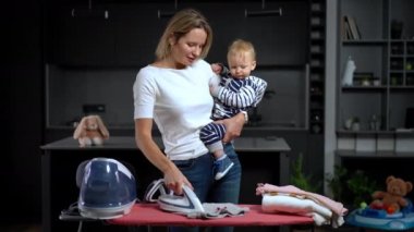 Portrait of happy confident mother ironing laundry talking with cute little baby. Caucasian beautiful woman doing housework chatting with infant daughter at home indoors. Maternity concept