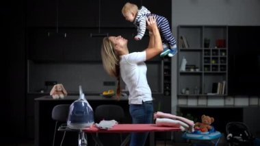 Side view cheerful woman raising baby girl up blowing on hair kissing child. Happy Caucasian mother playing with curios charming little daughter indoors at home. Leisure and love concept