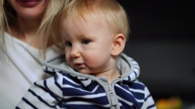 Close-up portrait of adorable curios blond baby girl with grey eyes looking around smiling as blurred unrecognizable woman talking. Happy charming Caucasian daughter in hands of mother indoors