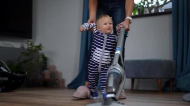 Cute cheerful infant girl walking helping woman vacuuming floor with vacuum cleaner. Charming smiling Caucasian daughter housekeeping with mother at home indoors. Live camera tracking shot