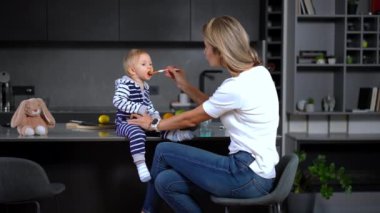 Cute Caucasian toddler sitting on tabletop as woman feeding baby with spoon. Portrait of pretty baby girl enjoying food as mother taking care of daughter indoors at home in kitchen