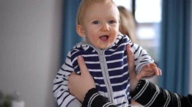 Cheerful adorable baby girl looking around smiling as woman playing with kid indoors. Portrait of pretty Caucasian toddler enjoying leisure with mother at home