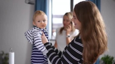 Side view curios cute baby girl looking at camera and looking around as young beautiful woman talking with kid. Blurred lady talking on phone at background