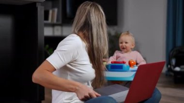 Positive confident woman messaging online on laptop playing with blurred infant in baby walker at background. Beautiful smart Caucasian mother working and taking care of baby. Multitasking maternity