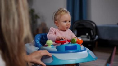 Portrait of baby girl looking at camera as woman riding baby walker in living room. Happy carefree Caucasian kid posing at home indoors playing with mother