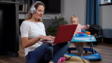 Positive busy Caucasian woman in headphones talking using video chat on laptop as curios infant in baby walker walking at background. Confident mother messaging online working as kid enjoying leisure