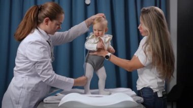 Curios baby girl playing with tape measure as doctor measuring height talking to happy mother. Caucasian charming infant on appointment in pediatric clinic with professional nurse chatting with woman