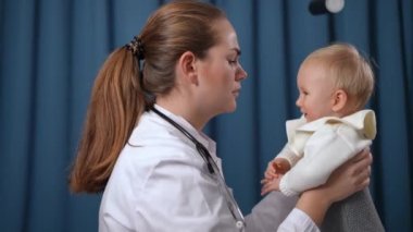 Side view positive pediatrician talking to baby girl cheering up little child. Beautiful Caucasian woman calming down dissatisfied infant in hospital indoors. Health care and infancy concept